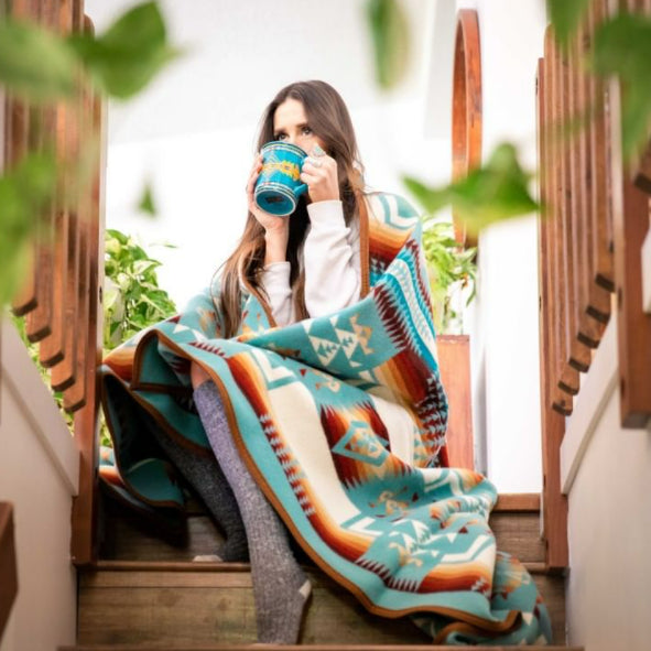 A woman sitting on stairs enjoying a cup of coffee from a turquoise Pendleton ceramin mug wrapped in a wool blanket that is colored aqua with brightly colored native design patterning