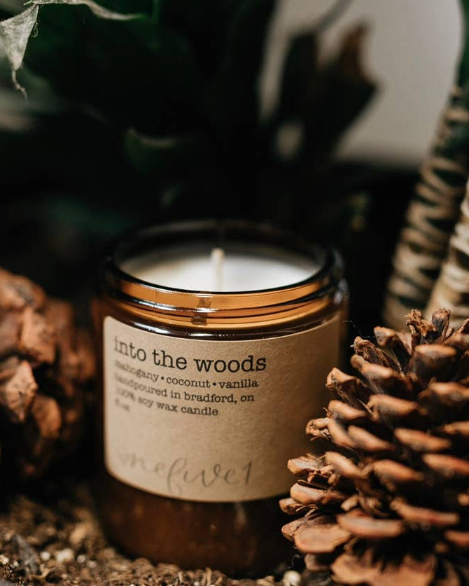 Candle labeled 'Into the Woods' surrounded by pine cones and plants