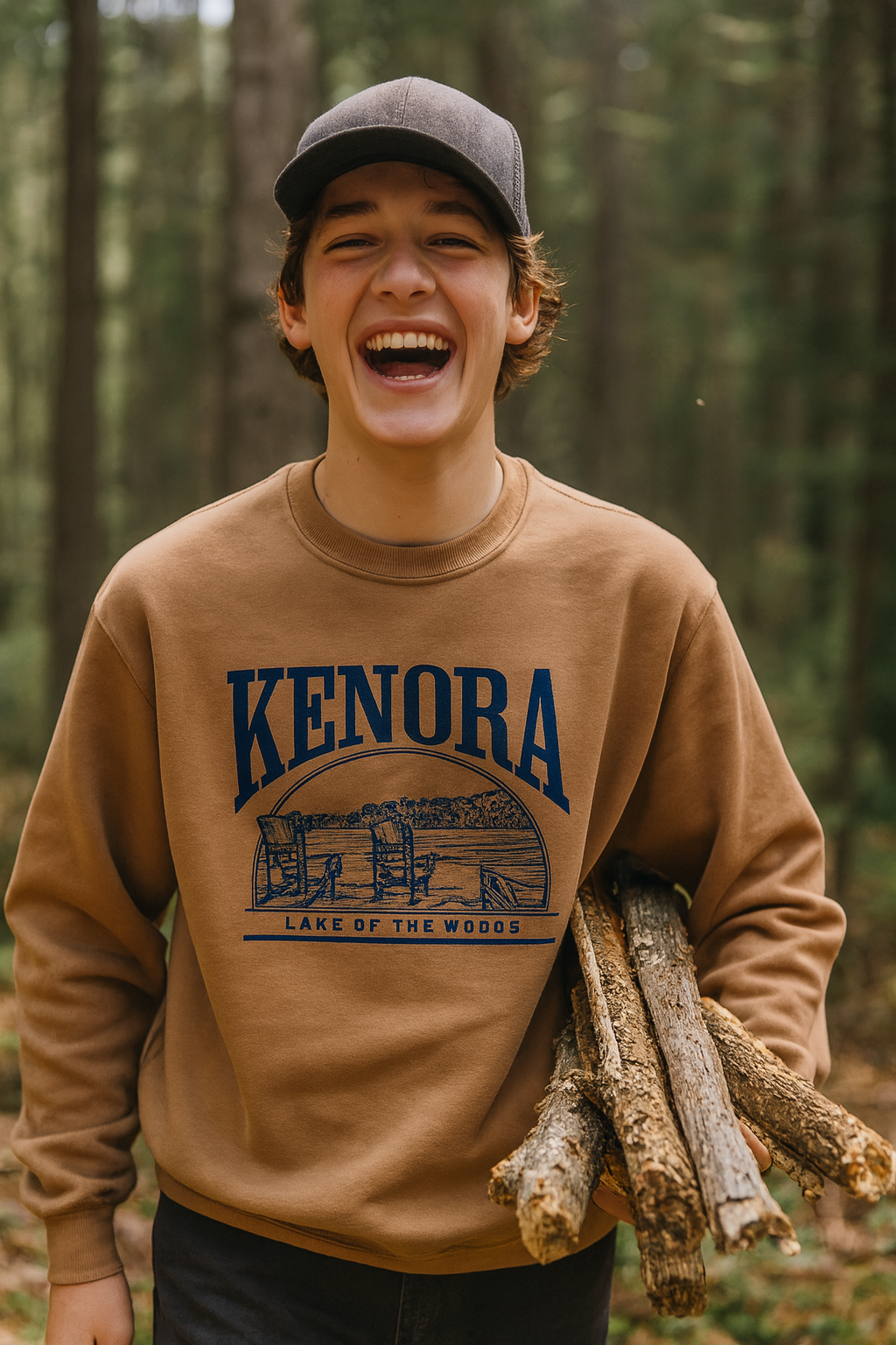 Teenage boy laughing joyfully in a forest while carrying firewood, wearing a camel-colored Kenora Lake of the Woods crewneck sweatshirt. The design on the sweatshirt features Adirondack chairs by the lake in navy blue. The image captures a fun, outdoor, adventurous vibe perfect for a souvenir or lifestyle brand.
