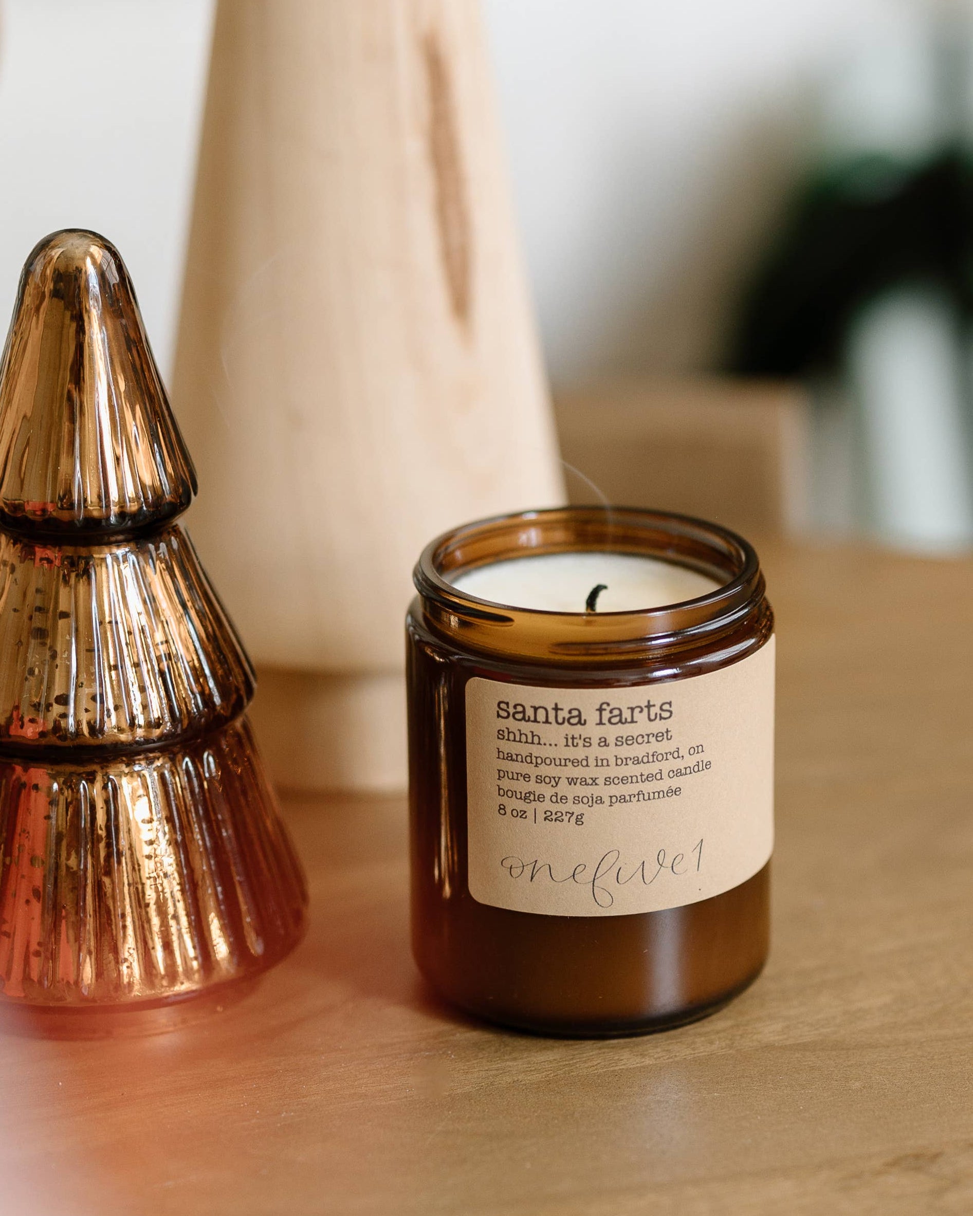 Candle labeled 'Santa Farts' next to a decorative tree on a wooden surface