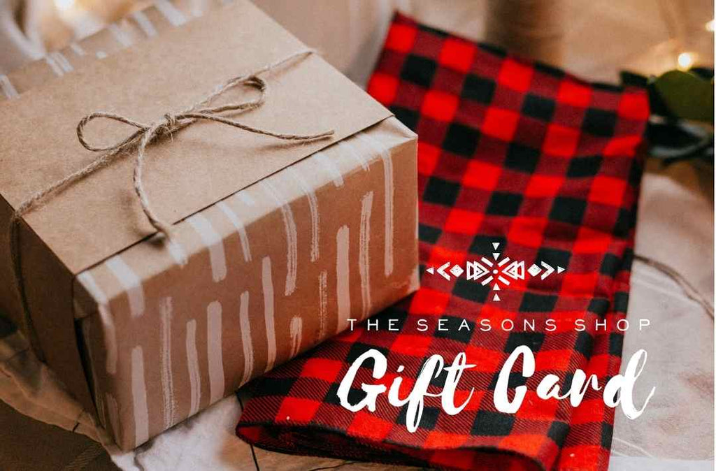 A beautifully wrapped gift on top of a red plaid blanket with words saying The Seasons Ship Gift Card