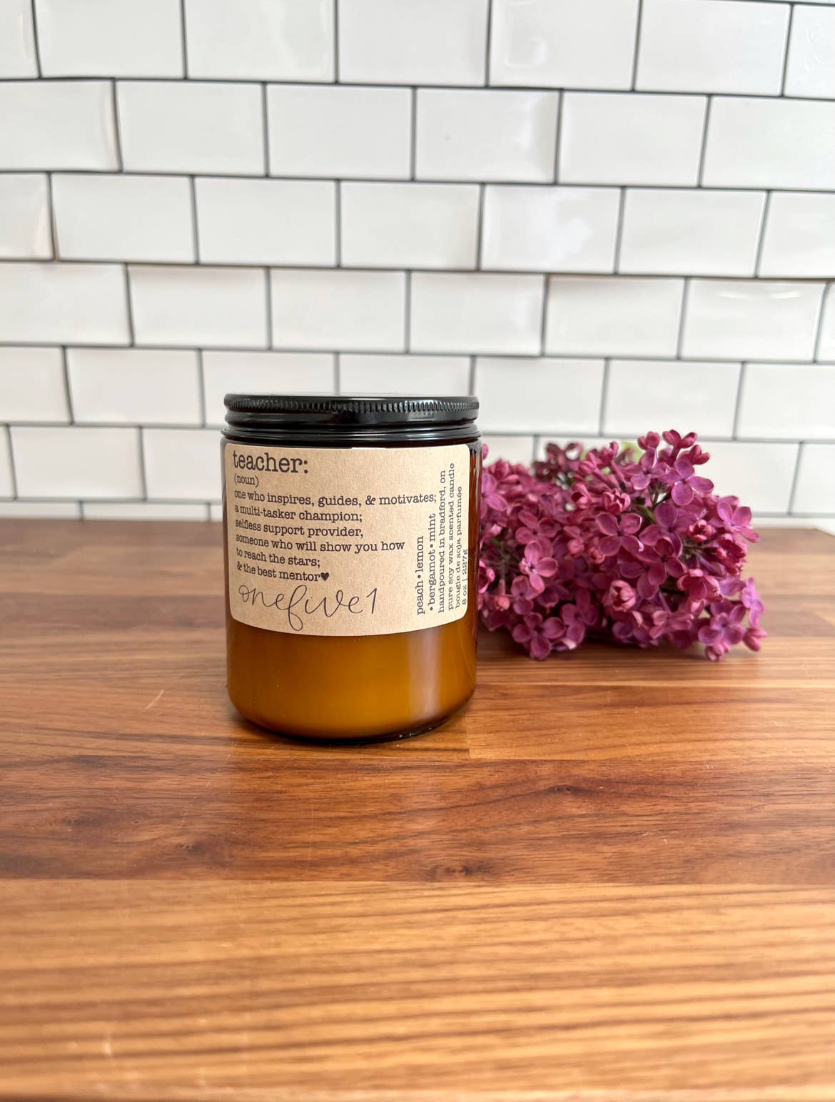 Candle with a label and purple flowers on a wooden surface against a tiled wall background
