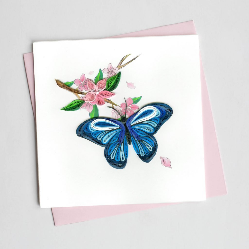 the front features a butterfly quilled in shades of blue resting on a branch with green leaves and pink flowers. 