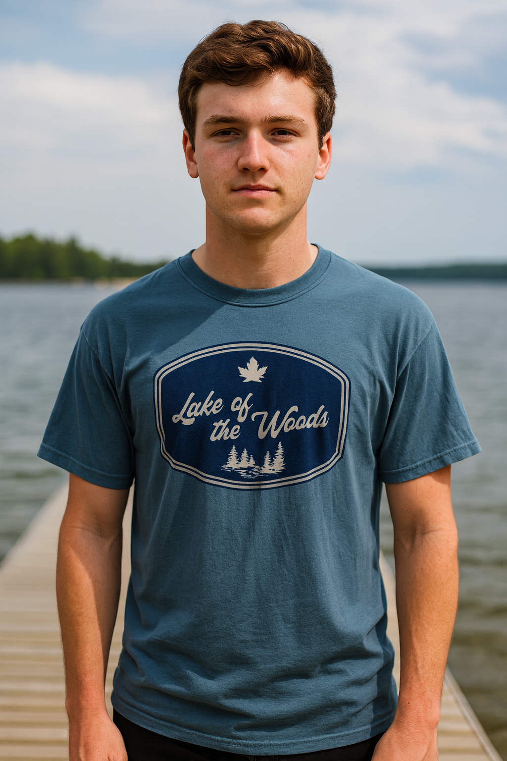 Young man standing on a dock by Lake of the Woods wearing a blue retro-style t-shirt with a navy logo featuring pine trees and a maple leaf. The shirt reads “Lake of the Woods” and is photographed outdoors with the lake and forest in the background