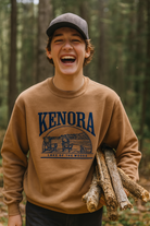 Teenage boy laughing joyfully in a forest while carrying firewood, wearing a camel-colored Kenora Lake of the Woods crewneck sweatshirt. The design on the sweatshirt features Adirondack chairs by the lake in navy blue. The image captures a fun, outdoor, adventurous vibe perfect for a souvenir or lifestyle brand.